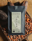 Tablette de chocolat noir Origines Chocolate Makers, emballée dans un sachet noir avec une étiquette verte indiquant 100 % de cacao provenant du Pérou, entourée de fèves de cacao et de cabosses sur un fond en toile de jute.