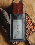 Tablette de chocolat noir Origines Chocolate Makers, emballée dans un sachet noir avec une étiquette bleue indiquant 70 % de cacao provenant de République Dominicaine, entourée de fèves de cacao et de cabosses sur un fond en toile de jute.