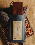Tablette de chocolat noir Origines Chocolate Makers, emballée dans un sachet noir avec une étiquette jaune indiquant 100 % de cacao provenant du Liberia, entourée de fèves de cacao et de cabosses sur un fond en toile de jute.