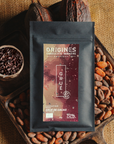 Tablette de chocolat noir au grué Origines Chocolate Makers, emballée dans un sachet noir avec une étiquette rouge indiquant 70 % de cacao provenant du Pérou, entourée de fèves de cacao, de cabosses et d'un bol contenant du grué de cacao sur un fond en toile de jute.