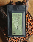 Tablette de chocolat noir Origines Chocolate Makers, emballée dans un sachet noir avec une étiquette verte indiquant 85 % de cacao provenant du Pérou, entourée de fèves de cacao et de cabosses sur un fond en toile de jute.