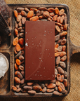 Tablette de chocolat noir à la fleur de sel du bassin d'Arcachon Origines Chocolate Makers, 70 % de cacao provenant du Pérou, entourée de fèves de cacao, de cabosses et d'un bol contenant du sel sur un fond en toile de jute.