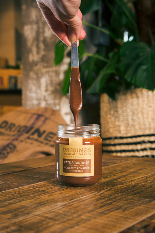 Pot de pâte à tartiner bio noisettes romaines et chocolat d'Origines Chocolate Makers, avec une cuillère soulevant la pâte, sur une table en bois dans un cadre chaleureux.