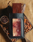 Tablette de chocolat noir au grué  Origines Chocolate Makers, emballée dans un sachet noir avec une étiquette rouge indiquant 70 % de cacao provenant du Pérou, entourée de fèves de cacao, de cabosses et d'un bol contenant du grué de cacao sur un fond en toile de jute.