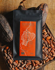 Tablette de chocolat noir Origines Chocolate Makers, emballée dans un sachet noir avec une étiquette orangeindiquant 70 % de cacao provenant d"Equateur, entourée de fèves de cacao et de cabosses sur un fond en toile de jute.