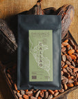 Tablette de chocolat noir Origines Chocolate Makers, emballée dans un sachet noir avec une étiquette verte indiquant 70 % de cacao provenant du Pérou, entourée de fèves de cacao et de cabosses sur un fond en toile de jute.