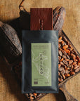Tablette de chocolat noir Origines Chocolate Makers, emballée dans un sachet noir avec une étiquette verte indiquant 70 % de cacao provenant du Pérou, entourée de fèves de cacao et de cabosses sur un fond en toile de jute.