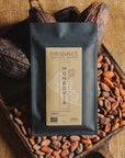 Tablette de chocolat noir Origines Chocolate Makers, emballée dans un sachet noir avec une étiquette jaune indiquant 100 % de cacao provenant du Liberia, entourée de fèves de cacao et de cabosses sur un fond en toile de jute.