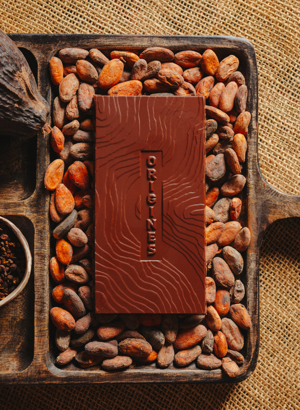 Tablette de chocolat noir au café  Origines Chocolate Makers 70 % de cacao provenant de Colombie, entourée de fèves de cacao, de cabosses et d'un bol contenant du café sur un fond en toile de jute.