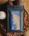 Tablette de chocolat noir à la fleur de sel du bassin d'Arcachon Origines Chocolate Makers, emballée dans un sachet noir avec une étiquette marron indiquant 70 % de cacao provenant du Pérou, entourée de fèves de cacao, de cabosses et d'un bol contenant du sel sur un fond en toile de jute.