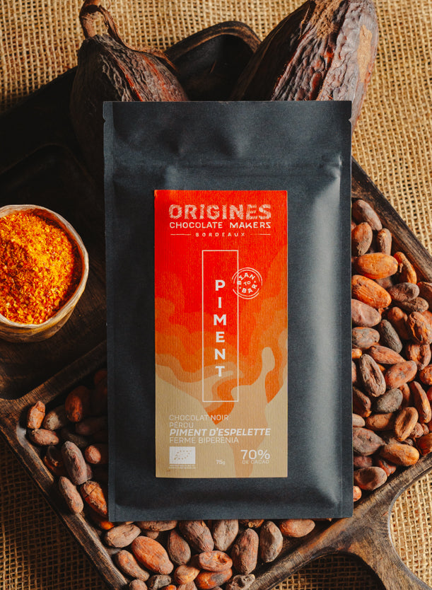 Tablette de chocolat noir au piment d'Espelette Origines Chocolate Makers, emballée dans un sachet noir avec une étiquette orange indiquant 70 % de cacao provenant du Pérou, entourée de fèves de cacao, de cabosses et d'un bol contenant du piment d'Espelette sur un fond en toile de jute.