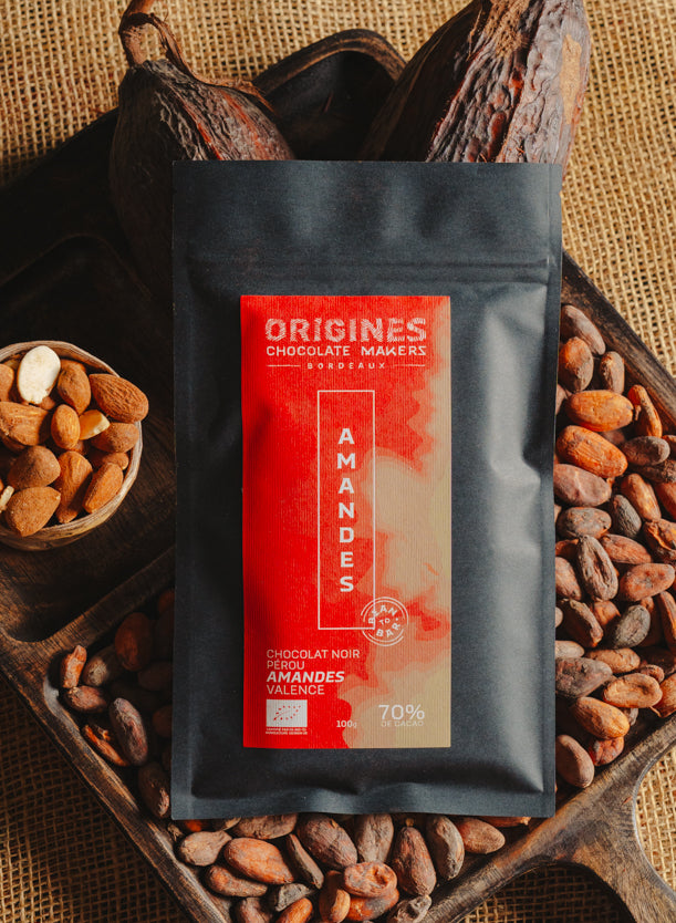 Tablette de chocolat noir au amandes de Valence Origines Chocolate Makers, emballée dans un sachet noir avec une étiquette rouge indiquant 70 % de cacao provenant du Pérou, entourée de fèves de cacao, de cabosses et d'un bol contenant des amandes sur un fond en toile de jute.