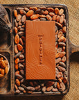 Tablette de chocolat au lait au amandes de Valence Origines Chocolate Makers,38 % de cacao de São Tomé entourée de fèves de cacao, de cabosses et d'un bol contenant des amandes sur un fond en toile de jute. 