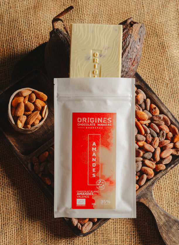 Tablette de chocolat blanc aux amandes de Valence Origines Chocolate Makers, emballée dans un sachet blanc avec une étiquette marron indiquant 35 % de cacao provenant du Pérou, entourée de fèves de cacao, de cabosses et d'un bol contenant des amandes sur un fond en toile de jute.