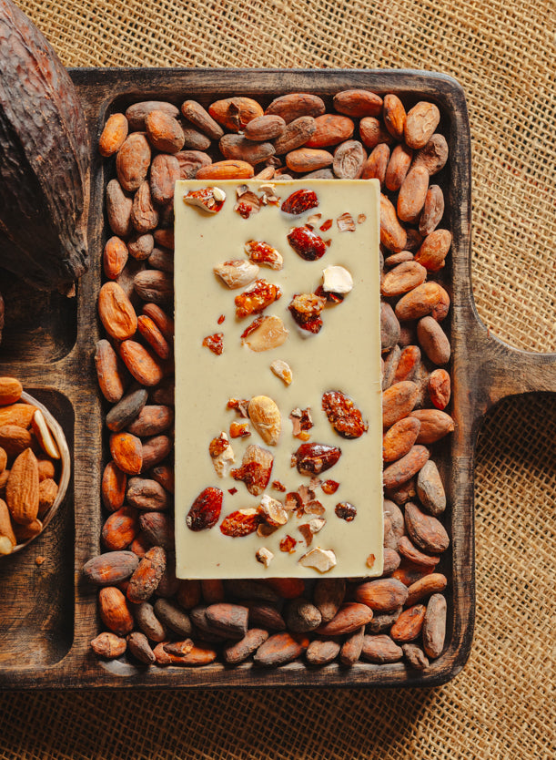 Dos d'une tablette de chocolat blanc aux amandes de Valence Origines Chocolate Makers 35 % de cacao provenant du Pérou, entourée de fèves de cacao, de cabosses et d'un bol contenant des amandes sur un fond en toile de jute.