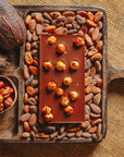 Dos d'une tablette de chocolat noir avec des noisettes de lot et garonne Origines Chocolate Makers 70 % de cacao provenant du Pérou, entourée de fèves de cacao, de cabosses et d'un bol contenant des noisettes sur un fond en toile de jute.
