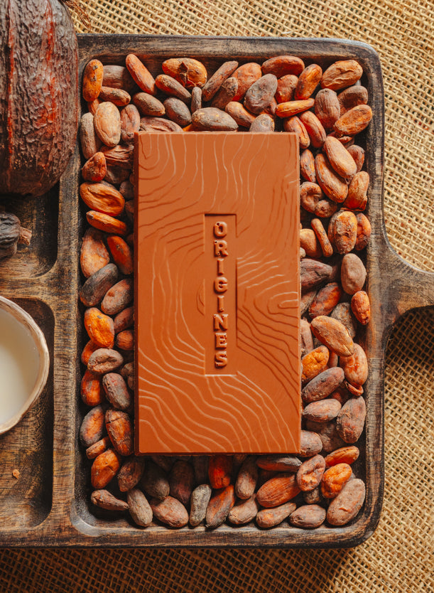 Tablette de chocolat au lait Origines Chocolate Makers, 38 % de cacao provenant de São Tomé, entourée de fèves de cacao, de cabosses et d'un bol contenant du lait sur un fond en toile de jute.