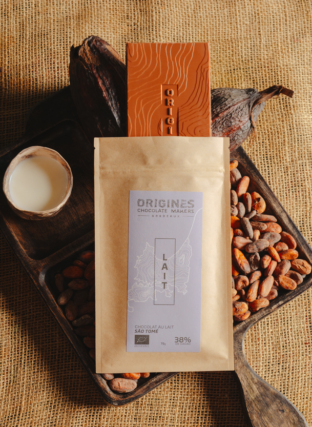 Tablette de chocolat au lait Origines Chocolate Makers, emballée dans un sachet kraft avec une étiquette violette indiquant 38 % de cacao provenant de São Tomé, entourée de fèves de cacao, de cabosses et d'un bol contenant du lait sur un fond en toile de jute.