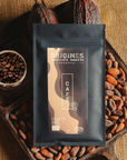 Tablette de chocolat noir au café  Origines Chocolate Makers, emballée dans un sachet noir avec une étiquette marron indiquant 70 % de cacao provenant de Colombie, entourée de fèves de cacao, de cabosses et d'un bol contenant du café sur un fond en toile de jute.