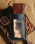 Tablette de chocolat noir au café  Origines Chocolate Makers, emballée dans un sachet noir avec une étiquette marron indiquant 70 % de cacao provenant de Colombie, entourée de fèves de cacao, de cabosses et d'un bol contenant du café sur un fond en toile de jute.