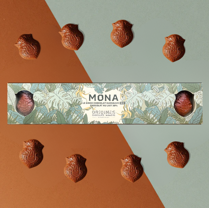 Chocolats en forme de singe 'Mona' de la marque Origines Chocolate Makers, disposés autour d'une boîte décorée de motifs végétaux, sur un fond bicolore marron et bleu clair