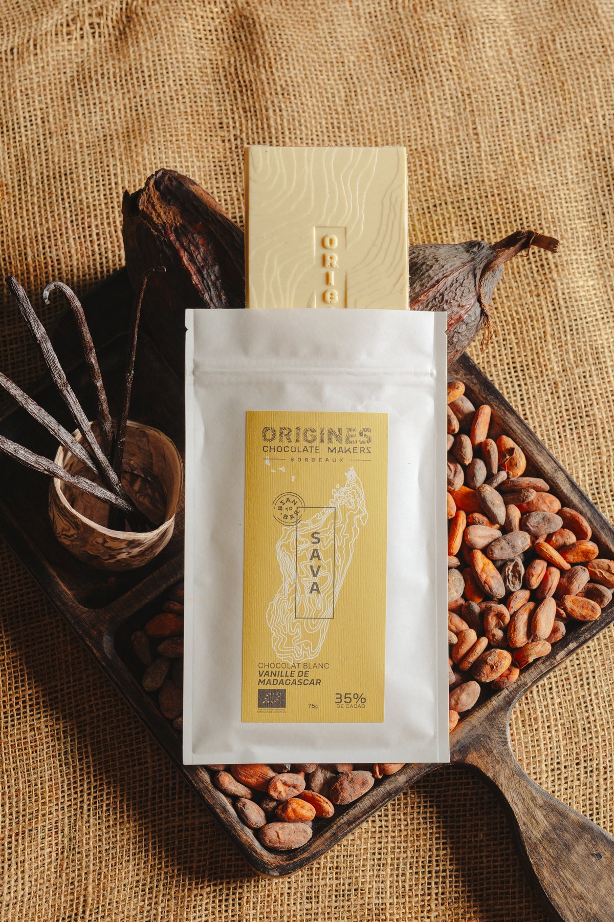 Tablette de chocolat blanc à la vanille Origines Chocolate Makers, emballée dans un sachet blanc avec une étiquette jaune indiquant 35% de cacao provenant de Madagascar, entourée de fèves de cacao, de gousses de vanille et de cabosses sur un fond en toile de jute.