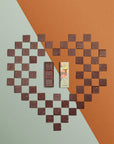 Une boîte rectangulaire de chocolats fins avec un toucan sur l'emballage est ouverte, révélant trois carrés de chocolat aux motifs variés, posée sur un cœur fait avec les carrés de chocolat et un fond géométrique dans les tons orange et vert.