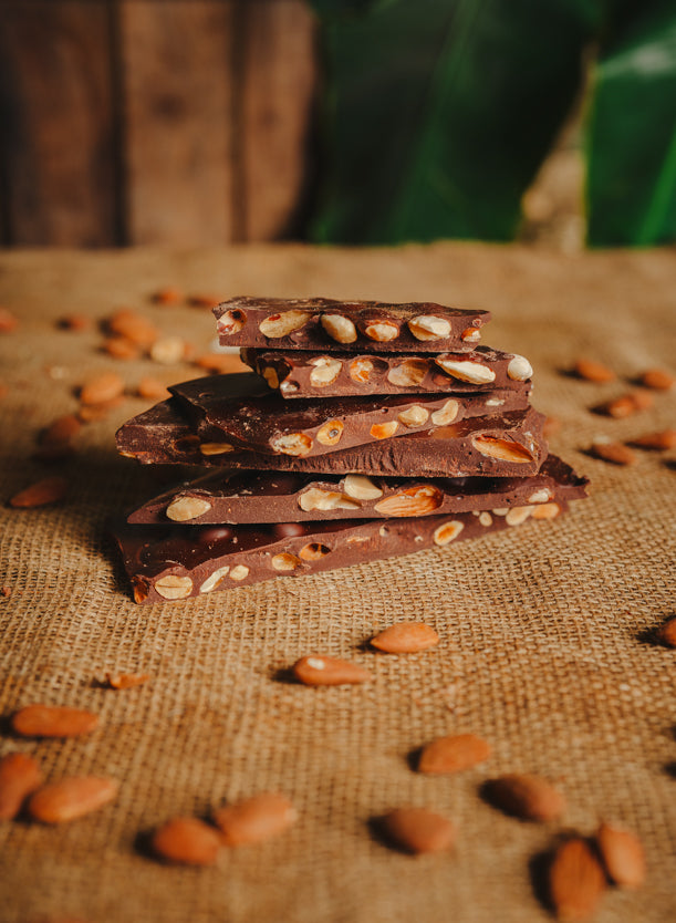 Morceaux de chocolat au lait aux amandes empilés sur une toile de jute, avec des amandes dispersées autour, en arrière-plan naturel.
