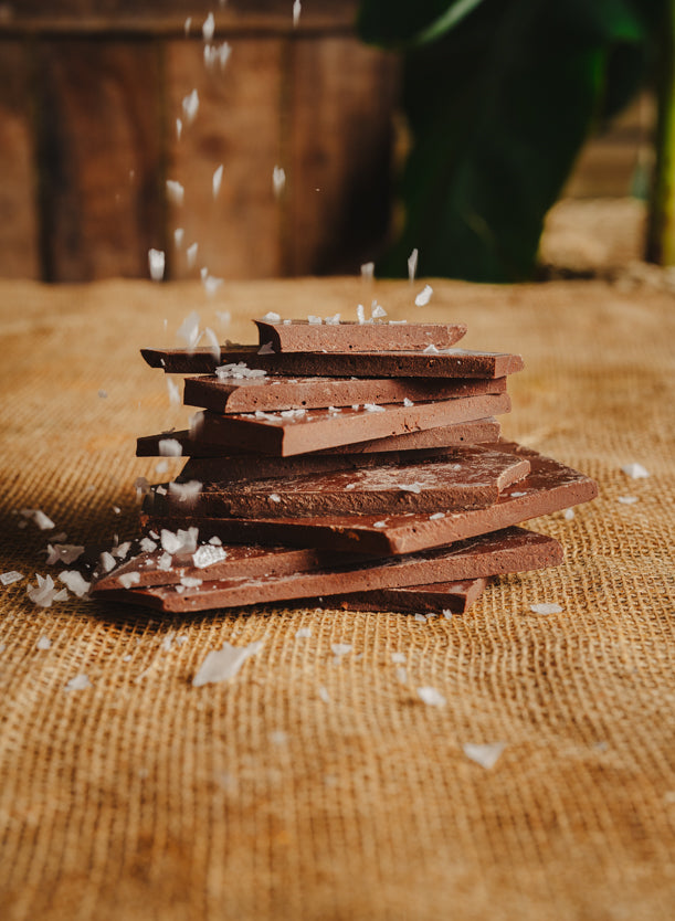 Morceaux de chocolat noir aux sel du bassin d'Arcachon empilés sur une toile de jute, avec du sel qui tombe sur le chocolat, en arrière-plan naturel.